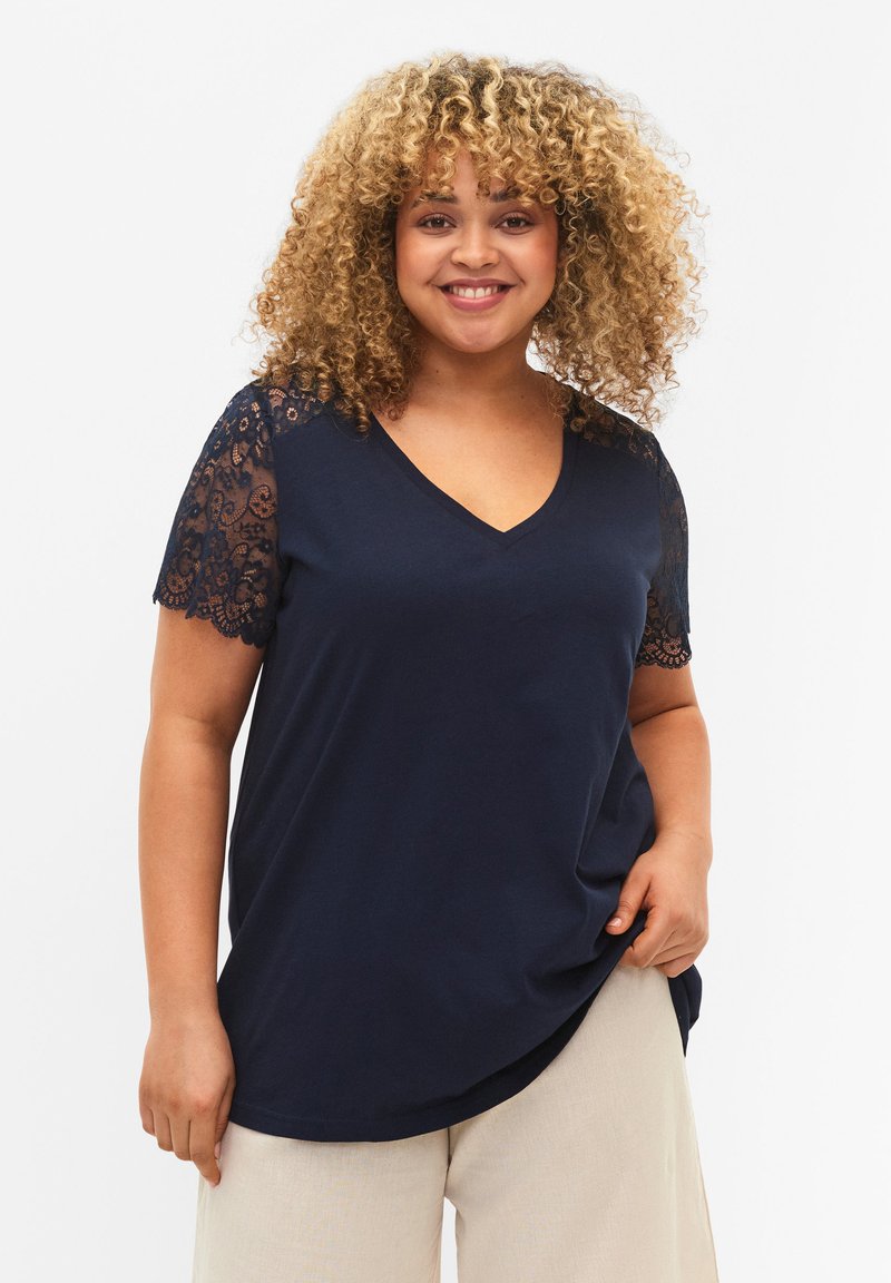 Camiseta navy de cuello en V con mangas cortas que presenta intrincados detalles de encaje negro en los hombros. La tela suave cae suavemente alrededor del cuerpo.