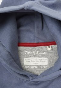 Harmaa puuvillaneule, jossa on sininen sisävuori ja punainen reuna. Tägissä on brändin nimi, koko ja orgaanisen sertifikaatin tiedot.