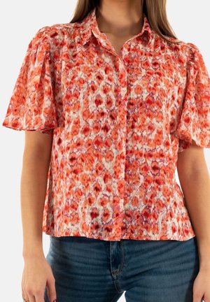 Femme portant un chemisier à manches courtes avec un motif abstrait orange et rouge, associé à un jean bleu foncé.