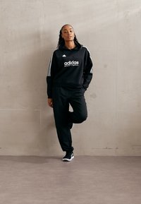 Melnā Adidas treniņtērpā ir kapucēta džemperis ar baltām svītrām un logo, kas saskaņots ar atbilstošām biksēm un melnām sporta kurpēm.