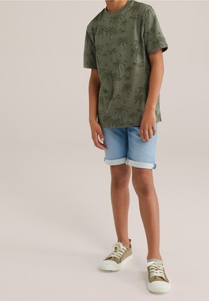 Child wearing an olive green palm tree print t-shirt, light blue rolled-up denim shorts, and beige sneakers with white laces, standing on a neutral floor.