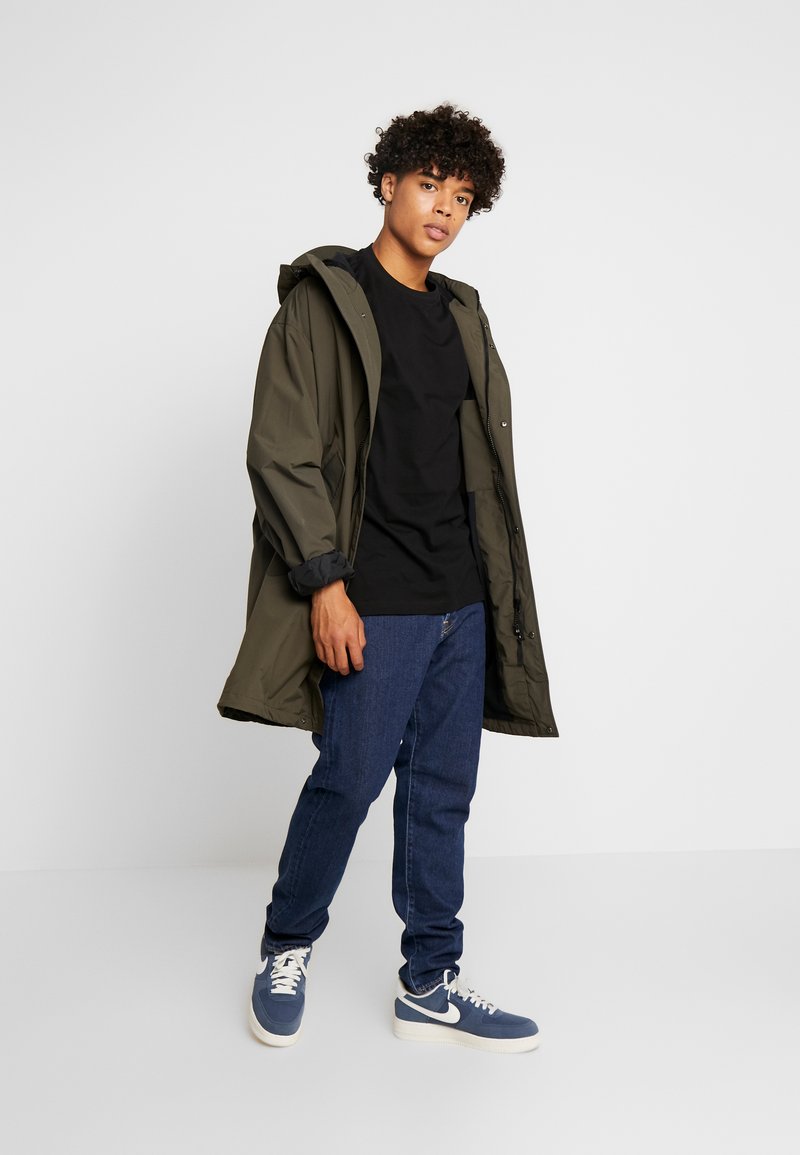 Olijfgroen parka met capuchon, zwarte t-shirt, blauwe denim jeans en blauwe sneakers. Bevat een relaxte pasvorm en drukknoopsluiting.