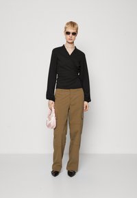 Top negru cu margini volănașe, pantaloni cargo bej cu buzunare și geantă din satin roz. Modelul poartă ochelari de soare și pantofi cu vârful ascuțit negri.
