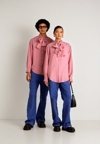 Duas modelos estão lado a lado, vestindo blusas rosa com laço combinando, calças azuis e sapatos pretos, uma delas segurando uma pequena mala preta.