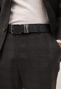 Schwarzer Ledergürtel mit einer glänzenden silbernen rechteckigen Schnalle, getragen zu einem grau karierten Anzug und einem hellen Strickpullover darunter.