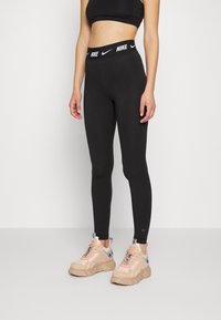 Svarta högmidjade leggings med ett tjockt midjeband som har Nike-logotypen. Bärs med ljusbeige och rosa sneakers med kontrasterande detaljer.