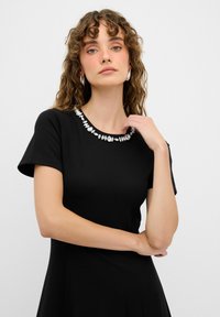 Schwarzes, kurzärmeliges Kleid mit rundem Ausschnitt, verziert mit dekorativen silbernen Applikationen. Glatter Stoff, taillierte Silhouette.