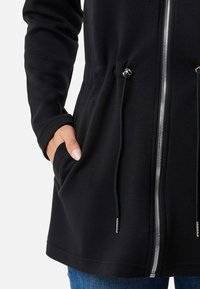Schwarze Zip-Jacke mit Kordelzug in der Taille, aus glattem Stoff gefertigt. Verfügt über Seitentaschen und silberne Reißverschlussdetails.