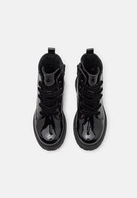 Un par de botas de tobillo de charol negro brillante con suelas gruesas y cordones negros, vistas desde arriba sobre un fondo blanco.