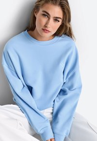 Gaiši zils džemperis ar brīvu piegriezumu, ribotiem apaļā kakla izgriezuma maliņām un nokarenām plecu līnijām. Krūšu rajonā ar mazu izšūtu logotipu.