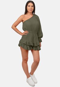 Rompers une épaule vert olive avec ourlet volanté, tissu texturé et taille élastique, associés à des baskets blanches.