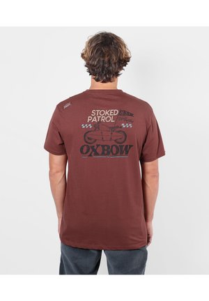 Homme aux cheveux ondulés debout de dos, portant un t-shirt marron avec un motif noir d'une planche de surf et le texte "Stoked Patrol Oxbow."