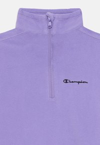 Pull en polaire lavande avec fermeture éclair quart de zip à l'avant et logo Champion brodé en noir sur la poitrine gauche.