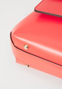 Coral pink handbag with a smooth, shiny texture, featuring gold hardware accents and contrasting stitching along the edges.
