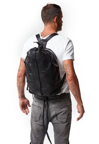 Mochila de cuero negro con forma redondeada, cierre de cremallera y correas ajustables, presentada en una persona que lleva una camiseta blanca y jeans grises.