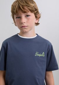 Ragazzino con capelli castani chiari e lentiggini, che indossa una maglietta blu scuro con il testo "The Scalpers" e un logo a forma di teschio sul petto.