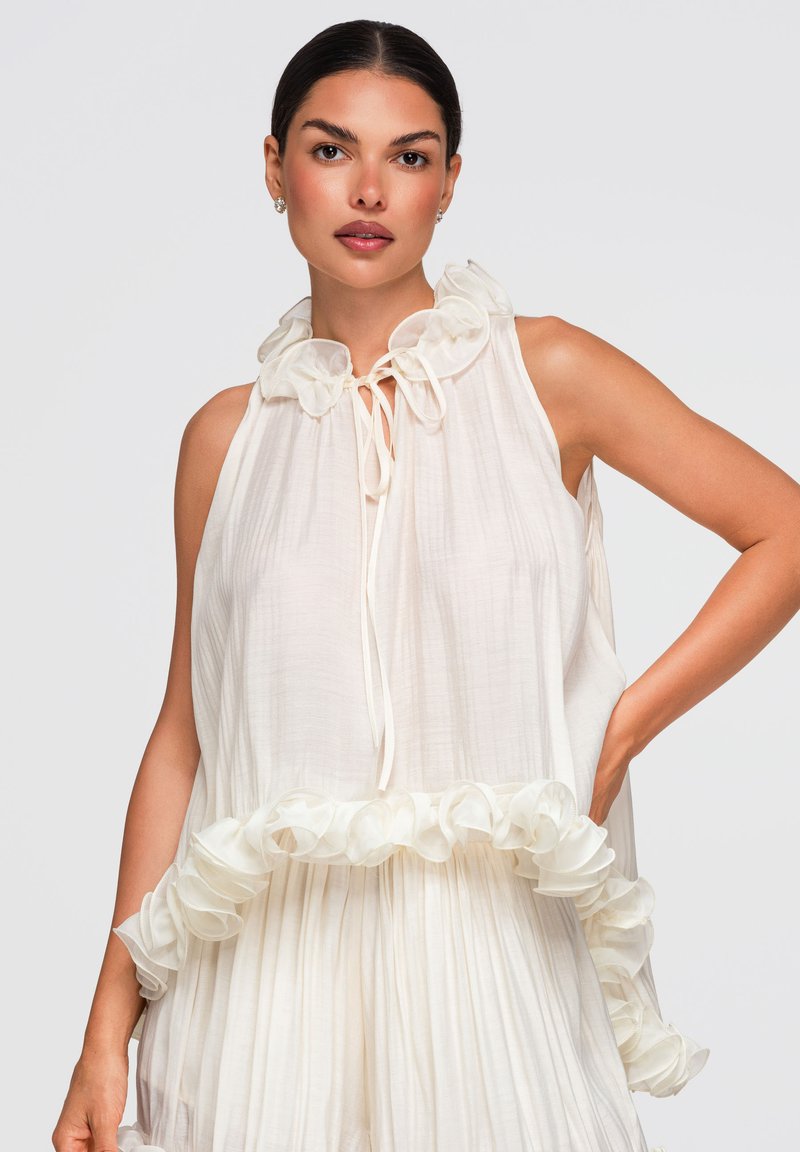 Femme aux cheveux foncés portant une robe blanche sans manches avec col et ourlet à volants, debout avec une main sur la hanche devant un fond uni.