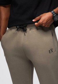 Zbliżenie na osobę w beżowych dresach z czarnym sznurkiem, zamkniętej kieszeni po boku oraz czarnym zegarku na lewym nadgarstku.