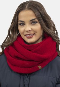 Red knitted infinity scarf made from soft yarn, featuring a textured pattern and a small branded tag, worn over a dark jacket.