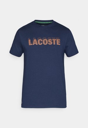 Camiseta de algodón azul marino de manga corta, con un gran logo texturizado de "LACOSTE" en color marrón contrastante en la parte frontal.