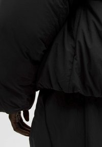 Dunjakke i sort, med en glat, skinnende stof, voluminøs silhuet og sammenkrøllet krave. Subtilt logo detajle synligt på siden.
