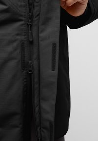 Cerrando la cremallera de una chaqueta negra y ajustando la solapa con tiras de Velcro sobre fondo blanco.