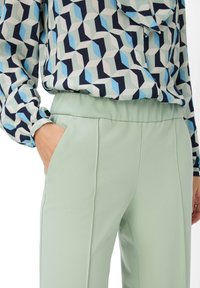 Gemusterte Bluse in Blau- und Marine-Tönen, kombiniert mit hellgrünen, glatten Hosen mit Seitentaschen und einem lockeren Schnitt.