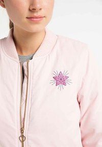 Roze bomberjack met een geribde kraag, gouden rits en een roze sterontwerp geborduurd op de borst met stralende lijnen.