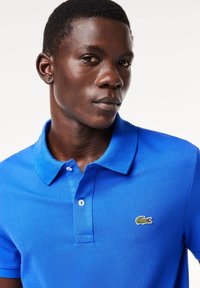 Camisa polo azul con una tela texturizada, dos botones en el cuello y un pequeño logo de cocodrilo verde y amarillo en el lado izquierdo del pecho.