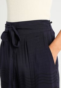 Mörkblå, högmidjade byxor med knytbälte, som har en slät textur och subtila vågmönster nära fållen. Sidoficka synlig.