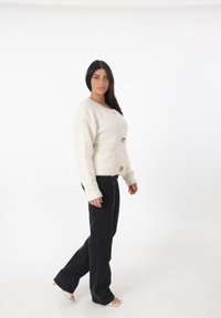 Cardigan bianco fluffy con tre grandi bottoni dorati, abbinato a jeans a flare neri. Il modello ha lunghi capelli scuri e si trova su uno sfondo bianco.