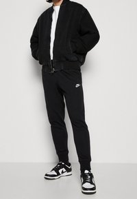 Svart bomberjacka med denimtextur, elastiska ärmar, bärs över en vit skjorta; matchad med svarta figurnära sweatpants med en vit logotyp.