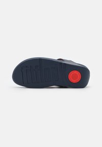 Svart gummisål med teksturert overflate, med "fitflop"-merke og en rød sirkulær detalj nær hælen.