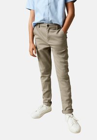 Beige színű, slim fit szabású chinos nadrág, gombos záródással és oldalzsebekkel, fehér tornacipővel párosítva. Világoskék rövid ujjú ing.