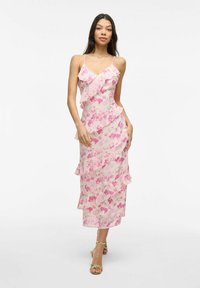 Kvinna som bär en rosa och vit blommig midiklänning med volanger och tunna spagettiremmar, kombinerat med guldhögklackade sandaler.
