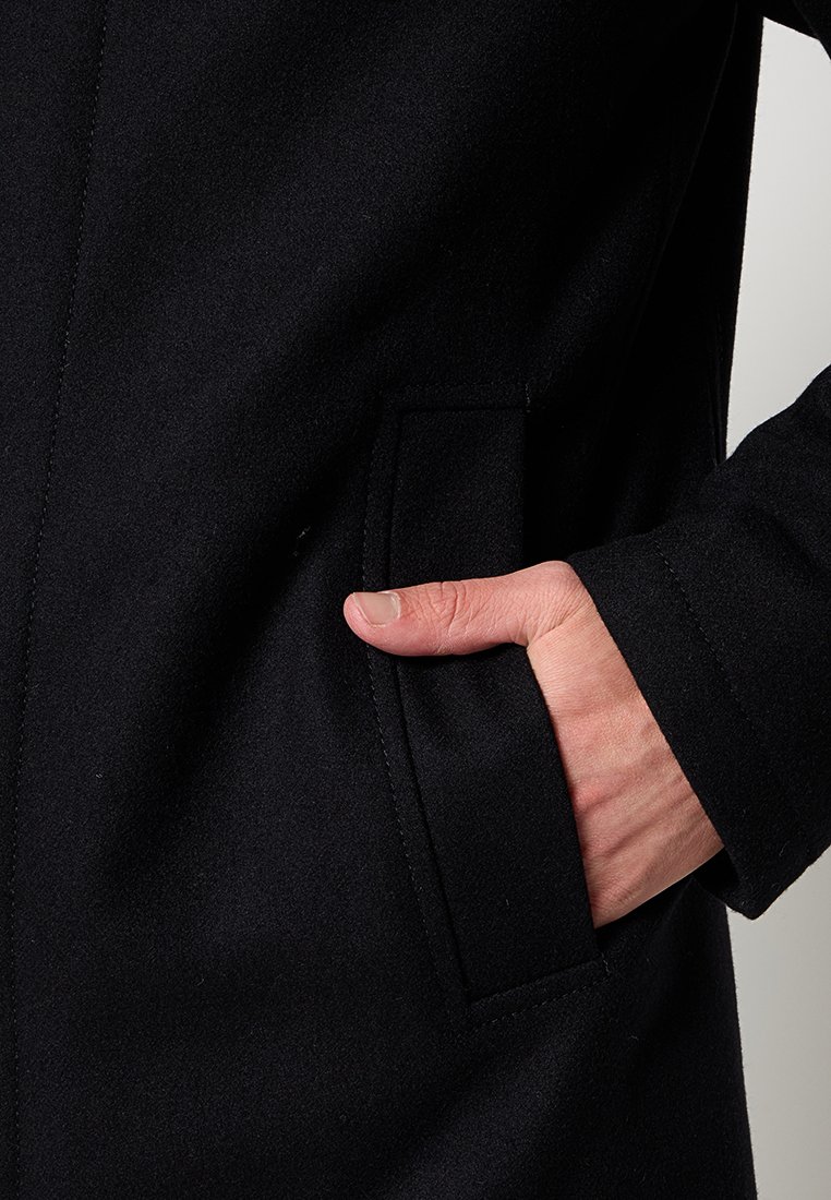Manteau en laine noire avec une poche sur le côté gauche, présenté dans un gros plan d'une main reposant à l'intérieur de la poche. Texture lisse, design simple.