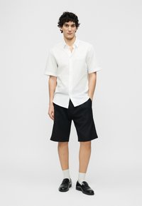 Άνδρας που φοράει λευκό κοντομάνικο πουκάμισο με κονκάρδα, μαύρα σορτς μέχρι το γόνατο, λευκές κάλτσες και μαύρα μοκασίνια, στέκεται με το ένα χέρι στην τσέπη.