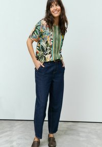 Chemise à manches courtes florale dans des tons de vert, orange et bleu, assortie à un pantalon en denim foncé. Le modèle se tient les mains dans les poches sur un fond clair.