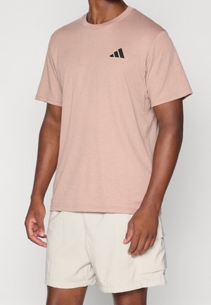Mand iført lysebrun t-shirt med sort logo og lyse beige shorts mod en ensfarvet baggrund.