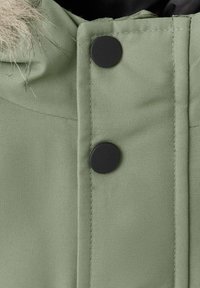 Tejido verde con un acabado texturizado, que presenta un botón a presión negro y un ribete de piel sintética, parte de la capucha de una chaqueta. Costuras horizontales visibles.