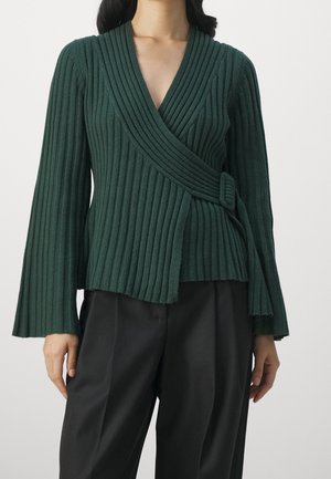 Femme portant un pull cache-cœur côtelé vert foncé à manches longues avec un détail à boucle, associé à un pantalon noir, sur un fond uni.