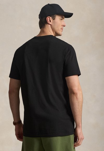 Άντρας που φοράει μαύρο καπέλο, μαύρο κοντομάνικο μπλουζάκι, πράσινο παντελόνι και καφέ ρολόι χειρός, στέκεται ελαφρώς στραμμένος δεξιά μπροστά σε μπεζ φόντο.