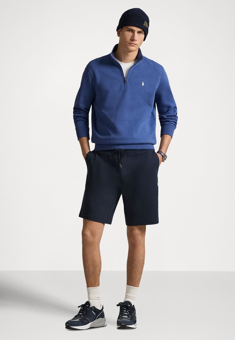 Giovane uomo con un maglione blu con cerniera a quarto, pantaloncini blu navy, berretto blu navy, calzini bianchi e scarpe da ginnastica blu navy, in piedi con le mani in tasca.