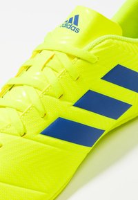 Scarpa sportiva Adidas gialla brillante con strisce e lacci blu, che mostra un primo piano della parte superiore e del logo sulla linguetta.