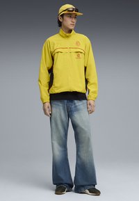 Gul zip-up jakke med røde detaljer og logo, parret med brede, falmede blå jeans og sorte sneakers. En matchende gul kasket fuldender looket.