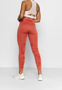 Czerwone legginsy w kamuflażu z teksturowanym pasem w talii, na którym znajduje się logo Puma. Materiał wydaje się elastyczny i dopasowany, wspierający ruch.