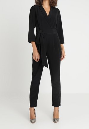 Femme portant une combinaison noire style cache-cœur avec des manches trois-quarts et une ceinture nouée sur le côté, accompagnée de talons pointus en peau de serpent.