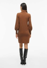 Abito in maglia marrone con colletto alto e maniche a palloncino lunghe, caratterizzato da una silhouette aderente e polsini a coste. Abbinato a stivali neri al ginocchio.