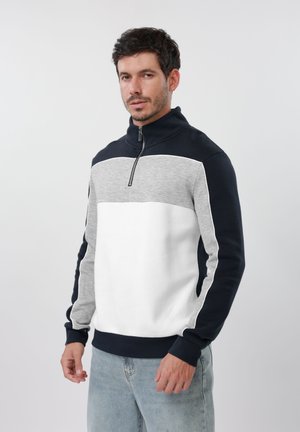 Hombre que lleva un pulóver con bloques de color con cuello negro, parte superior del pecho gris, torso blanco y vaqueros de lavado claro, de pie contra un fondo liso.