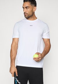 Λευκό αθλητικό t-shirt με ραμμένη υφή και κοντά μανίκια, με το λογότυπο "BOSS". Το μοντέλο κρατάει μια νεον κίτρινη μπάλα τένις και ρακέτα.